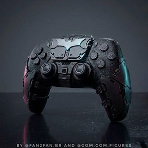 Op de afbeelding: Een zwarte gamecontroller met een getextureerde grip en een centrale plaat met een zilverkleurig vleermuisvormig embleem. Het apparaat heeft metaalachtig ogende zijpanelen met mechanische details en de tekst BY @FAN2FAN.BR AND @GOM.GOM.FIGURES.