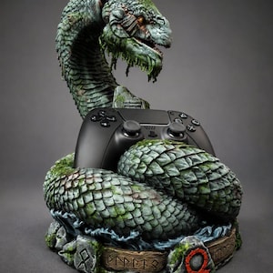 Puede incluir: Un mando de videojuego negro que descansa entre los anillos de una detallada escultura de serpiente en tonos verdes y grises. La base presenta texturas de piedra con inscripciones rúnicas y un símbolo circular rojo con una línea.