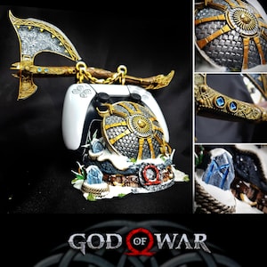 May include: A gaming controller stand featuring a replica axe and shield with gold and silver metallic finishes. The base is decorated with snow, rope, and blue crystal accents. The text God of War is displayed at the bottom of the image.