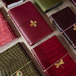 Pode incluir: Uma coleção de diários retangulares com várias texturas e cores, incluindo vermelho profundo, verde-oliva e bordô. Cada diário é preso com uma faixa elástica adornada com pingentes de metal dourado em forma de flores, libélulas e sóis.