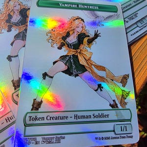 May include: A rectangular holographic trading card titled Vampire Huntress, featuring an illustration of a blonde character in a dark green dress with a yellow sash, white stockings, and brown boots, accompanied by a white owl, with text Token Creature - Human Soldier.