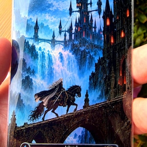 May include: A rectangular trading card titled Island from the Vampyr Land Series, featuring a rider on a black horse crossing a stone bridge toward a dark gothic castle with glowing red windows, set against a backdrop of blue waterfalls.