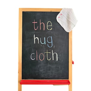 Puede incluir: Una pizarra de madera con una bandeja roja muestra el texto "the hug cloth" escrito con tiza de colores. Un paño de tela blanco, texturizado y cuadrado con dos pequeños corazones rosas está colocado sobre la esquina superior derecha.