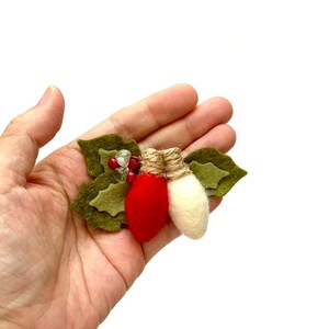 Op de afbeelding: Een kerstversiering van vilt met twee lampjes, een rode en een witte, met groene hulstbladeren en rode bessen.
