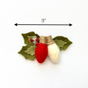 Op de afbeelding: Een kerstversiering van vilt met twee lampjes, een rood en een wit, met een accent van jute touw. De lampjes zijn omgeven door groene vilt hulstbladeren en rode en transparante kralen. De versiering is 7,6 cm breed.