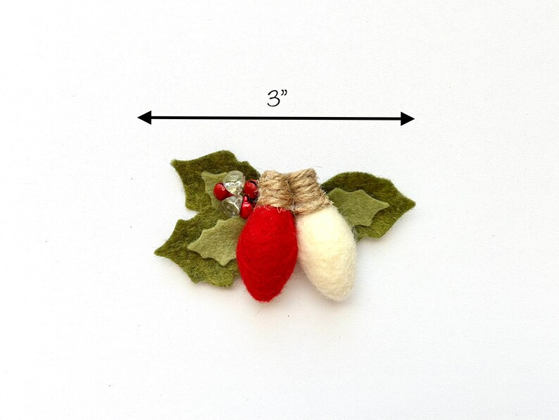 Op de afbeelding: Een kerstversiering van vilt met twee lampjes, een rood en een wit, met een accent van jute touw. De lampjes zijn omgeven door groene vilt hulstbladeren en rode en transparante kralen. De versiering is 7,6 cm breed.