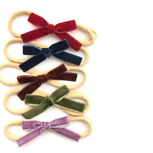 May include: Five velvet hair bows in different colors, attached to nude elastic headbands. The colors are burgundy, navy blue, brown, forest green, and lavender.