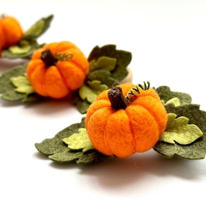 May include: Three felt pumpkins with green felt leaves. The pumpkins are orange and have a brown stem.