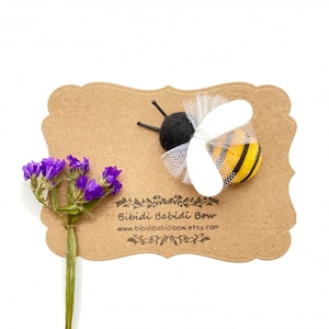 May include: A yellow and black felt bee with white tulle wings on a brown card with the text "Bibbidi Babbidi Bow" and the website address "www.bibbidi babbidi bow.etsy.com".