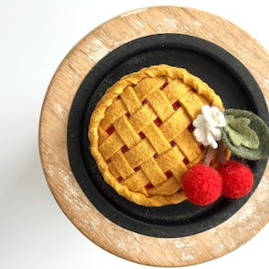 May include: A felt pie with a lattice crust, red filling, and cherry accents. The pie sits on a black plate, which is set on a wooden base. The cherries are red with green leaves and a white flower.