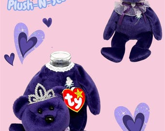 Op de afbeelding: Een paarse pluche beer met een zilveren kroon en een witte kanten kraag met een paars lint. De beer heeft een zwarte neus en een rood hartvormig Ty-label. De tekst Plush-N-Stuff Buddy staat in blauw schrift naast afbeeldingen van paarse harten.