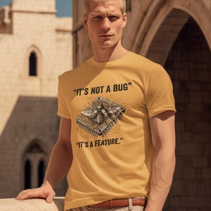 May include: A mustard yellow short-sleeve t-shirt featuring a graphic of a mechanical insect integrated into a computer circuit board. The text above the graphic reads IT'S NOT A BUG and the text below the graphic reads IT'S A FEATURE.