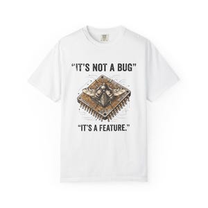 May include: A white short-sleeve t-shirt featuring a graphic of a brown computer processor chip with a large insect resting on top. The text above the graphic reads IT'S NOT A BUG and the text below the graphic reads IT'S A FEATURE.