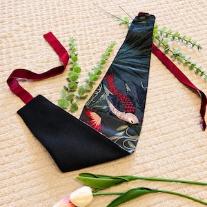 Peut inclure: Un bandana triangulaire pour animal avec un tissu sombre orné d'un motif d'oiseaux rouges et blancs parmi un feuillage vert. L'accessoire présente une face en tissu noir uni et de longs rubans rouge foncé pour l'attacher.