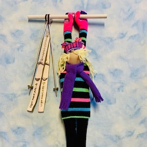 May include: A handmade doll with blonde yarn hair, wearing a purple and black striped shirt, a purple scarf, and pink and white striped socks. The doll is hanging from a white bar with two pairs of ski poles hanging below.