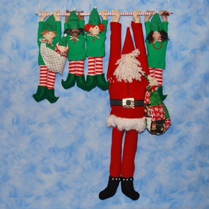 May include: A Santa Claus doll hangs from a clothesline with five elf dolls. The Santa doll is wearing a red suit with a white fur trim and a black belt. The elf dolls are wearing green and red outfits with white fur trim.