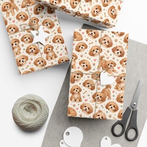 May include: Wrapping paper featuring a repeating pattern of light brown curly-haired puppies, small paw prints, pink hearts, and beige bones on a cream background. The paper is tied with twine and includes a white star-shaped or heart-shaped gift tag.