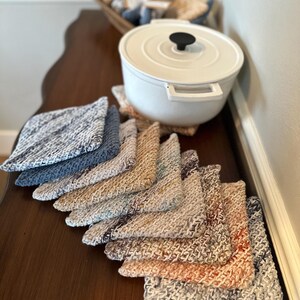 Könnte beinhalten: Ein Stapel quadratischer, gehäkelter Topflappen aus Baumwolle in verschiedenen melierten Farben, darunter Blau-, Beige-, Creme- und Rosttöne, fächerförmig angeordnet neben einem weißen gusseisernen Topf mit schwarzem Knauf auf dem Deckel.