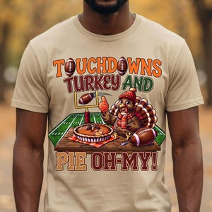 May include: A beige t-shirt featuring a graphic of a turkey wearing a red knit hat and scarf, standing on a wooden table next to a pie and a football. The text reads TOUCHDOWNS TURKEY AND PIE OH-MY! with footballs and a goalpost in the background.