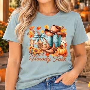 May include: A light blue t-shirt featuring a graphic print of a pumpkin with a plaid bow, a brown cowboy hat, and turquoise cowboy boots surrounded by autumn leaves and sunflowers, with the text Howdy Fall in brown script.