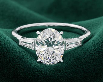 May include: A silver-coloured ring featuring a large, central oval-cut clear gemstone held by four prongs, flanked on each side by a single, smaller, elongated baguette-cut clear gemstone set into the polished metal band.