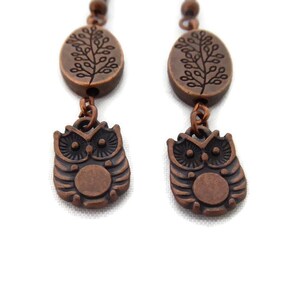 Puede incluir: Un par de pendientes de color cobre. Cada pendiente presenta un pequeño dije ovalado con un diseño de árbol, conectado a un colgante de búho detallado. Los colgantes de búho tienen ojos grandes y una barriga circular. Los pendientes cuelgan de una cadena de cuentas.