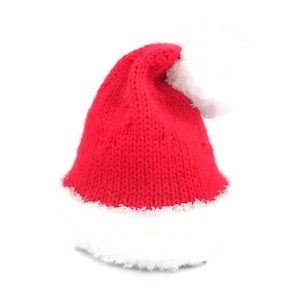 Puede incluir: Un gorro de punto rojo brillante que presenta un borde blanco grueso y difuso en la base y un pompón blanco grande y suave unido a la parte superior. El gorro está construido con un material de hilo texturizado con un patrón de punto visible.