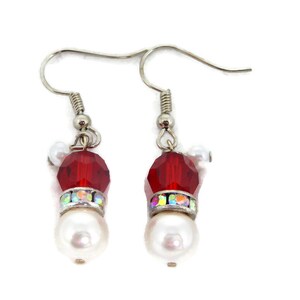 Puede incluir: Un par de pendientes festivos con un diseño de gorro de Papá Noel. Cada pendiente tiene una cuenta roja facetada, una banda de pedrería iridiscente y una perla blanca grande. Los pendientes tienen ganchos para las orejas de color plateado.