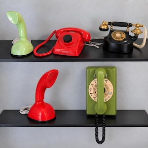 Può includere: Una collezione di cinque telefoni esposti su due ripiani neri. Il ripiano superiore presenta una cornetta verde chiaro, un telefono da scrivania rosso con cavo a spirale e un telefono nero in stile antico. Il ripiano inferiore contiene una cornetta rossa.