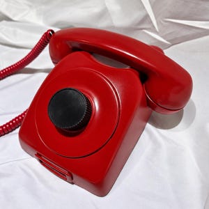 May include: A bright red landline telephone featuring a smooth, glossy finish and a matching coiled cord. The device has a prominent, circular black dial or knob centred on the main body, with a traditional handset resting securely on top.
