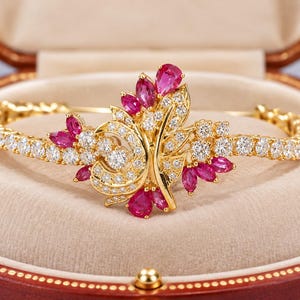 Puede incluir: Una pulsera de oro con un diseño floral central engastado con rubíes rojos en talla pera y diamantes blancos redondos. La banda consiste en una fila continua de diamantes blancos redondos, creando una pieza de joyería simétrica.