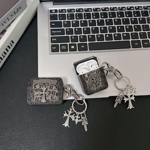Puede incluir: Dos fundas protectoras negras para auriculares inalámbricos, con un patrón de letras góticas oscuras y emblemas de cruz de metal plateado. Cada funda incluye un llavero de metal plateado con dijes colgantes en forma de cruz y daga.