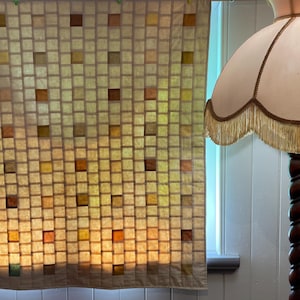 May include: A square quilt featuring a grid of small, light-coloured fabric squares interspersed with occasional brown, yellow, and orange squares. Beside it stands a floor lamp with a twisted wooden base and a cream-coloured shade with fringe.