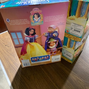 Può includere: Una scatola di giocattoli rettangolare in cartone con Biancaneve e i sette nani della Disney. La parte anteriore mostra un'immagine di una bambola di Biancaneve e due bambole di nani, Cucciolo e Eolo, con testo tra cui 'Disney Classics' e 'Snow White'.