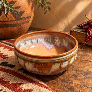 May include: A round ceramic bowl featuring a reactive glaze with blended tones of warm orange, deep brown, and creamy white. The interior and exterior surfaces display a smooth, glossy finish with vertical drip patterns cascading down the sides.