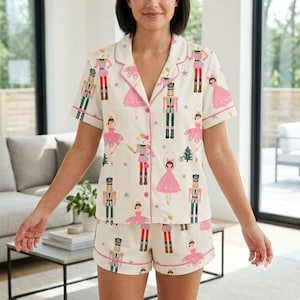 May include: A cream-colored short-sleeve pajama set featuring a repeating pattern of nutcrackers and ballerinas in pink, green, and gold. The button-down shirt has a notched collar with pink piping, and the matching shorts feature an elastic waistband.