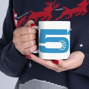 Peut inclure: Un mug en céramique blanche arborant un grand chiffre 51 bleu clair imprimé en gras sur le côté. Le design intègre une icône de trèfle à quatre feuilles blanc stylisé dans la barre horizontale du chiffre cinq, créant un graphisme épuré.