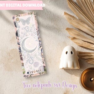 May include: A rectangular notepad featuring a celestial design with a central crescent moon, butterflies, and floral motifs in shades of purple and grey. The bottom edge displays a lunar phase cycle. The text reads Two notepads 3x8 Design.