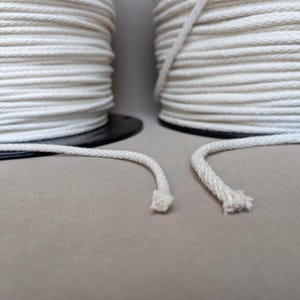 Puede incluir: Dos grandes carretes de cordón trenzado de color blanco roto, cada uno enrollado firmemente sobre una base circular negra. La cuerda gruesa y texturizada se extiende desde los carretes, con los extremos deshilachados visibles en primer plano.