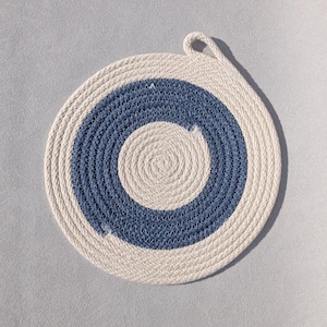 May include: A circular trivet constructed from coiled rope featuring a central cream-coloured circle, a middle ring of blue rope, and an outer border of cream-coloured rope. A small loop of rope is attached to the top edge for hanging purposes.