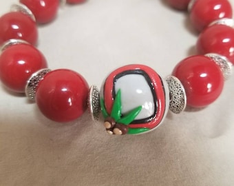 May include: Red beaded bracelet with a single white bead featuring a green leaf design and brown accents.