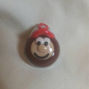 May include: A brown, cartoon-like character with a smiling face, large eyes, and a red hat. The character is round and has a light tan face with a dark brown outline. The hat is red with a white band.