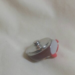 May include: A miniature, decorative pot with a silver lid and a dark red body. The pot has a white and red handle. This small kitchenware item is likely a collectible or a toy.
