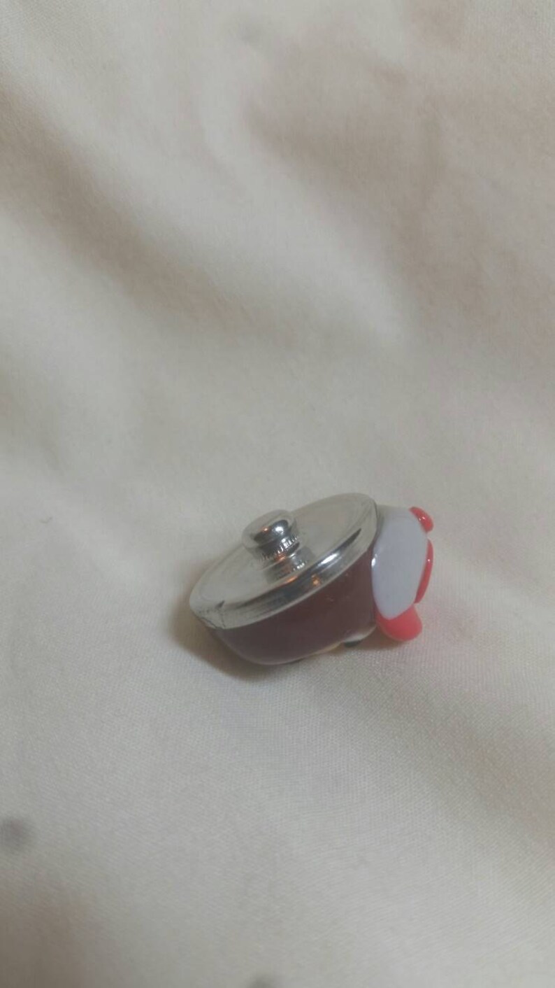 May include: A miniature, decorative pot with a silver lid and a dark red body. The pot has a white and red handle. This small kitchenware item is likely a collectible or a toy.