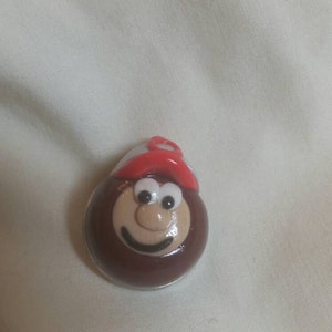 May include: A round, smiling cartoon character with a brown body, tan face, and a red and white hat. The character has white eyes and a black smile. The item is likely a pin or a decorative item.