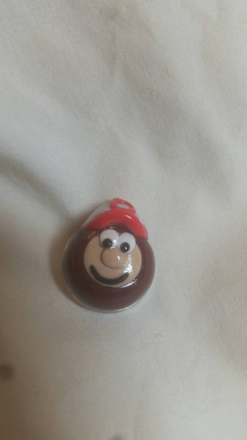May include: A round, smiling cartoon character with a brown body, tan face, and a red and white hat. The character has white eyes and a black smile. The item is likely a pin or a decorative item.