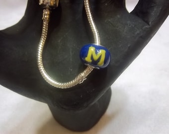 May include: A silver charm bracelet with a blue and yellow charm featuring the letter "M".