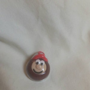 May include: A brown, oval-shaped figurine with a smiling face and a red hat. The figurine has a white and red hat and a smiling face. The figurine is on a white surface.