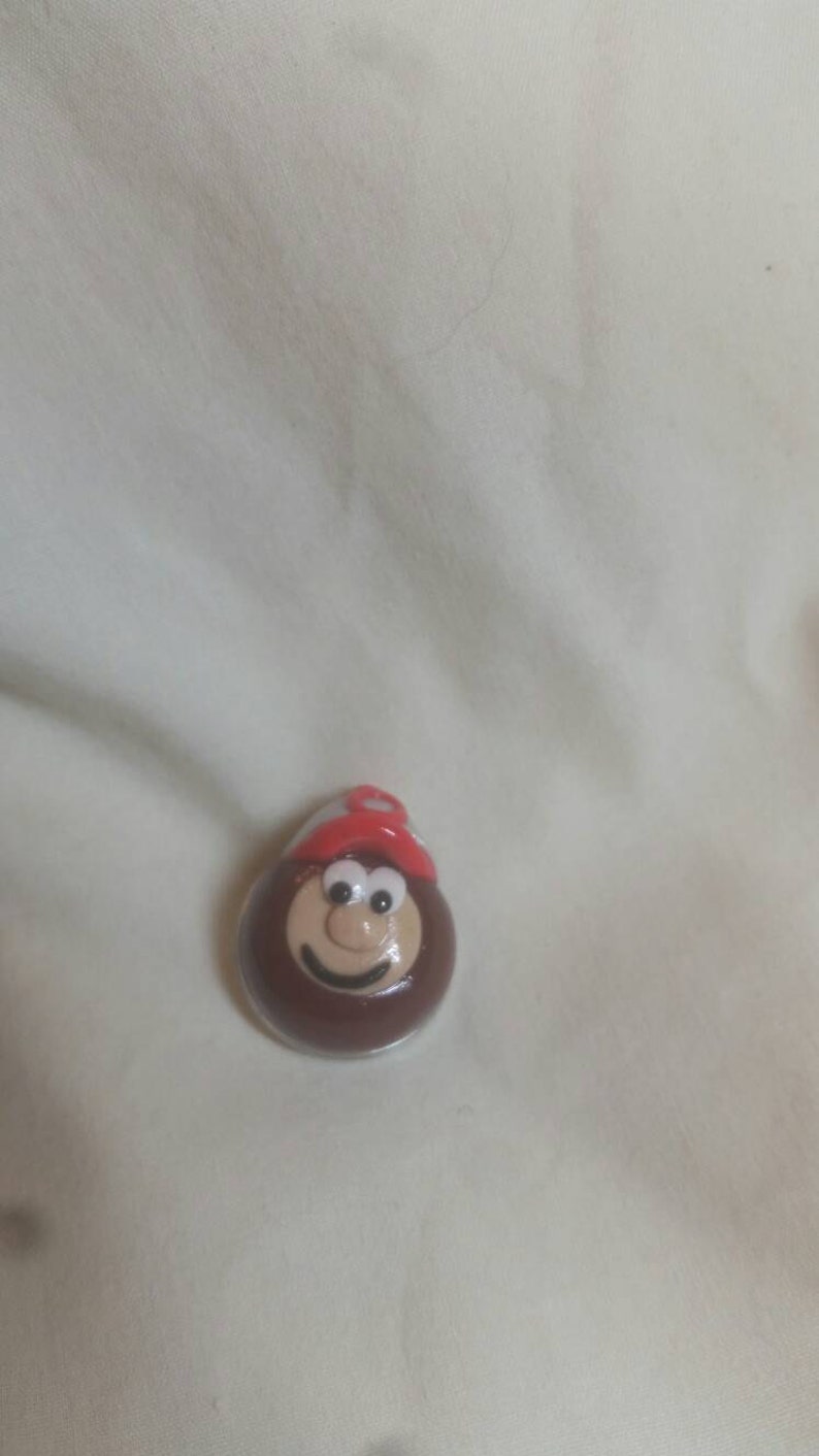 May include: A brown, oval-shaped figurine with a smiling face and a red hat. The figurine has a white and red hat and a smiling face. The figurine is on a white surface.