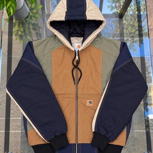 May include: A color-blocked hooded jacket featuring a tan center panel, olive green upper chest sections, and navy blue sleeves. The jacket includes a full-length front zipper, black ribbed cuffs, and a Carhartt brand patch on the lower front pocket.